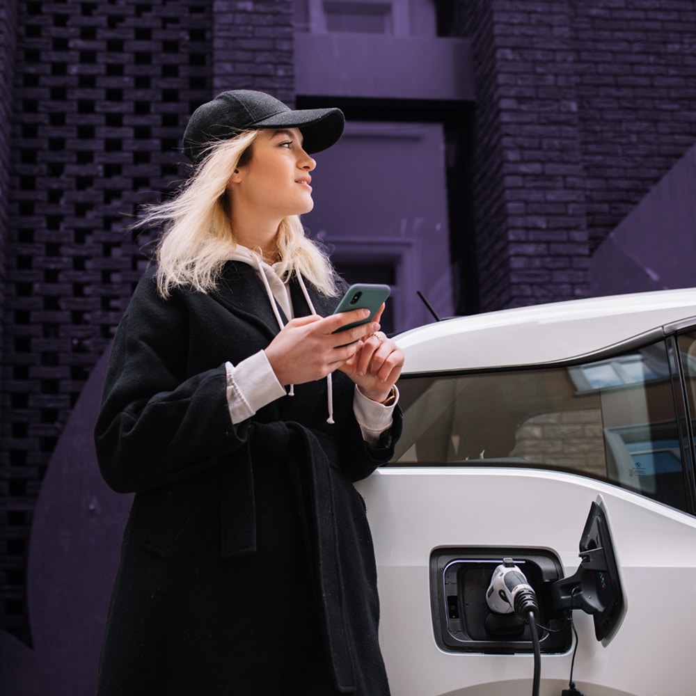 A woman stands in front of an electric vehicle at the charging station, making a satisfied face as it charges successfully. She taps playfully on her smartphone.